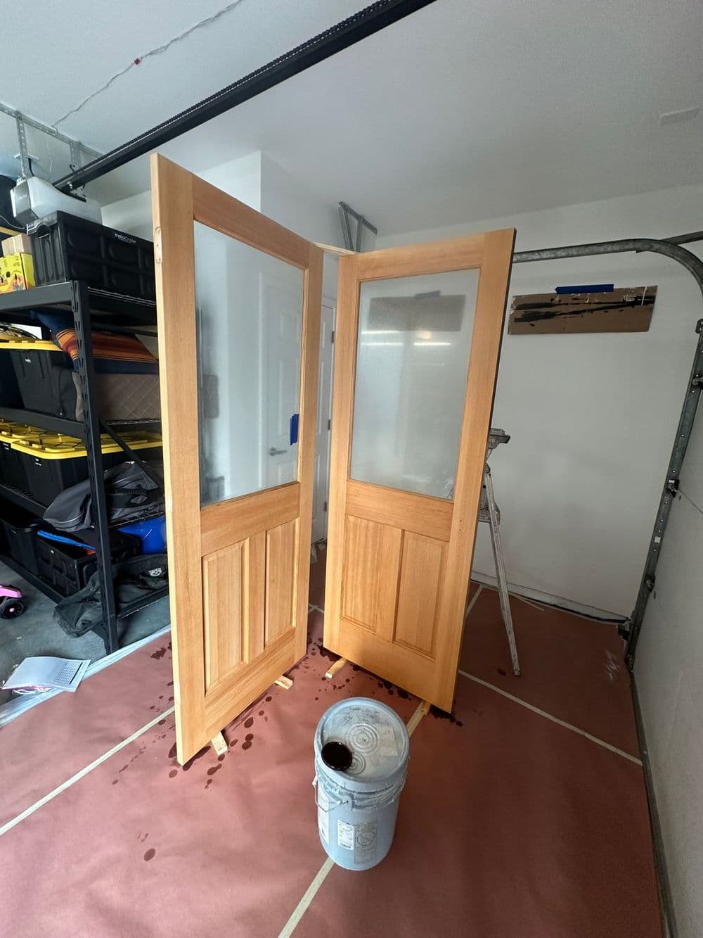 Wooden doors with glass panels leaning in a garage, next to a paint bucket on the floor.
