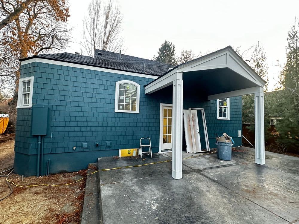 Blue shingle house exterior with porch and unfinished landscaping. Construction materials nearby.