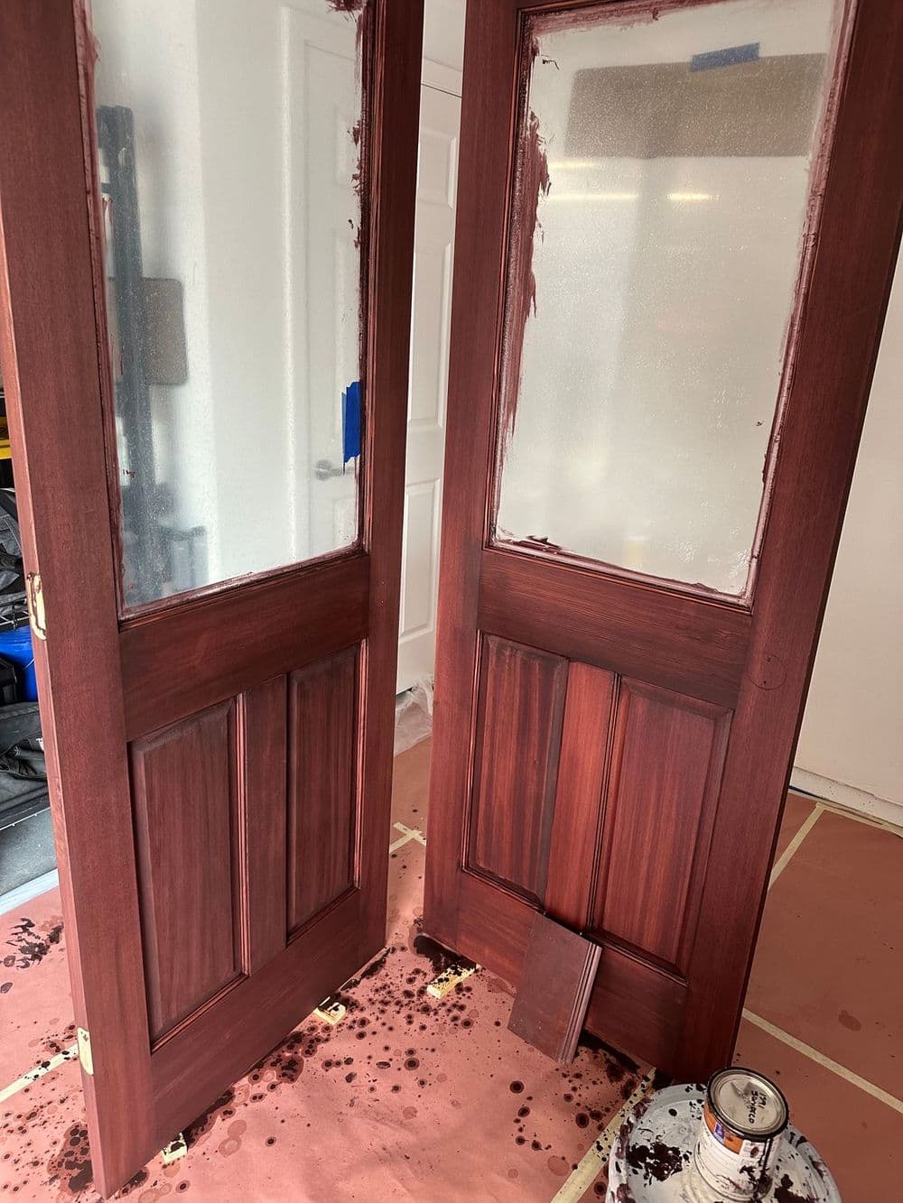 Two wooden doors being painted, with brown paint dripped on the floor and clear panels.