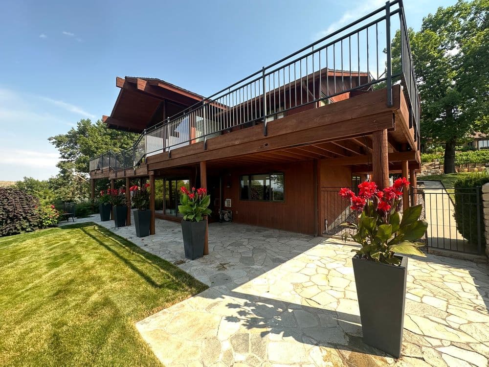Modern wooden house with large balcony, surrounded by greenery and red flowers in planters.