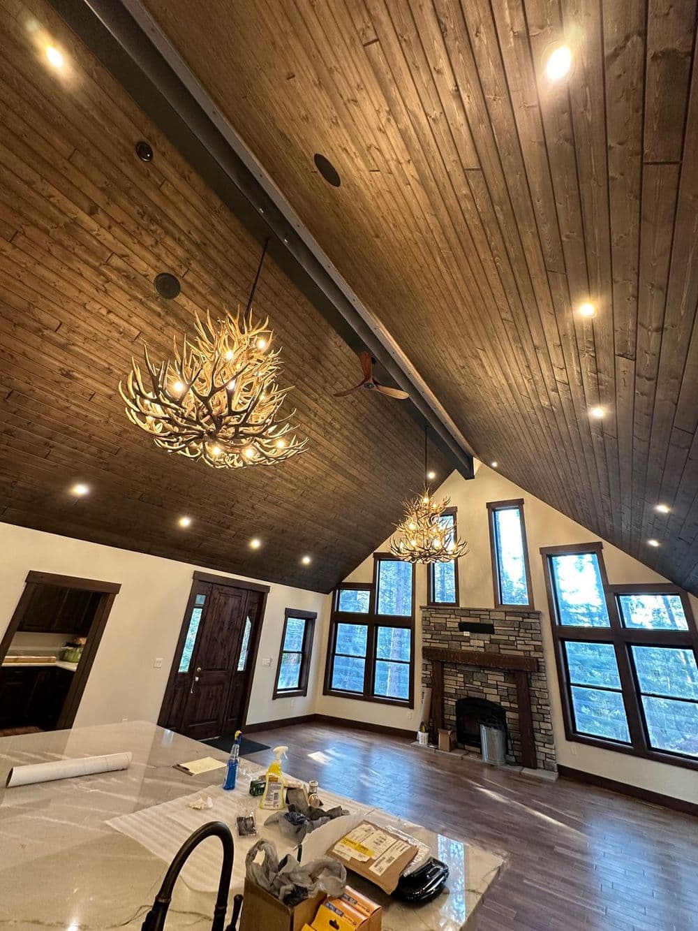 Modern rustic living room with wooden ceiling, stone fireplace, and unique antler chandeliers.