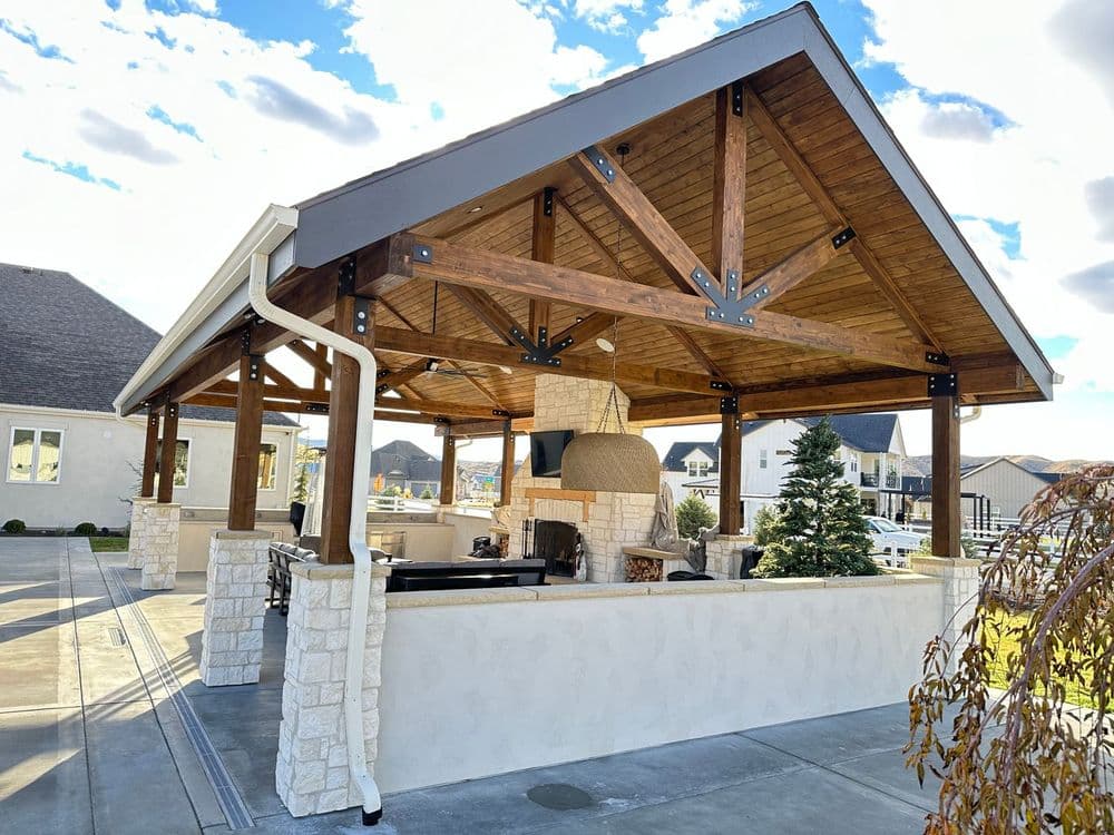 Outdoor pavilion with wooden beams, stone fireplace, and cozy seating area under a blue sky.