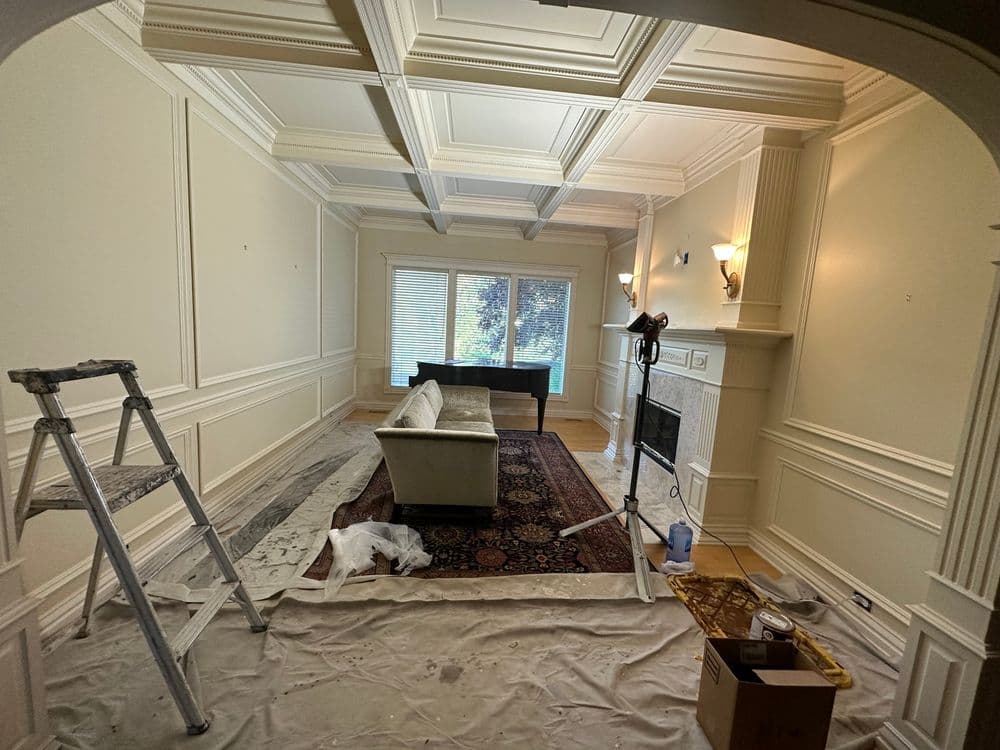Living room with white walls, coffered ceiling, and a piano, in the midst of painting renovation.