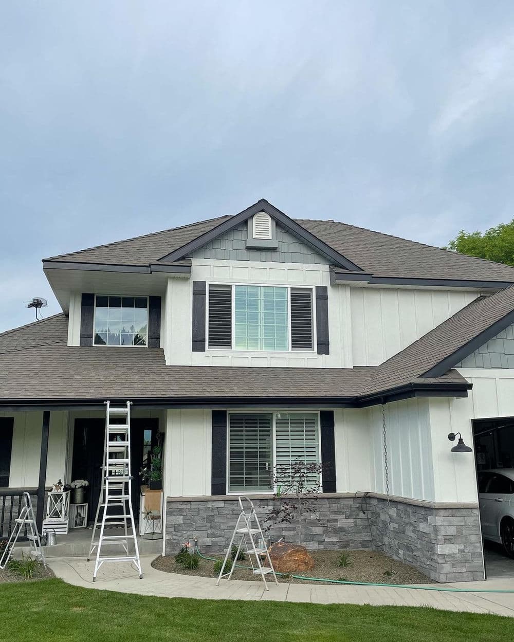 Two-story house with shutters, ladders, and stone foundation under a cloudy sky.