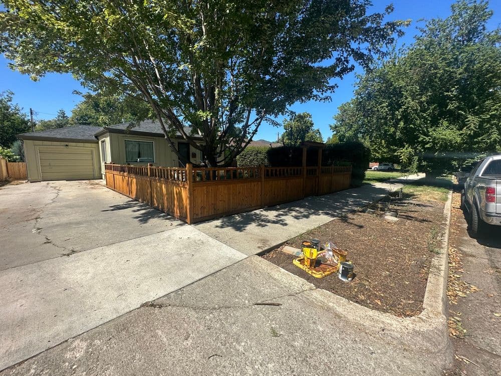 Residential house with wooden fence, large tree, and driveway on a sunny day.