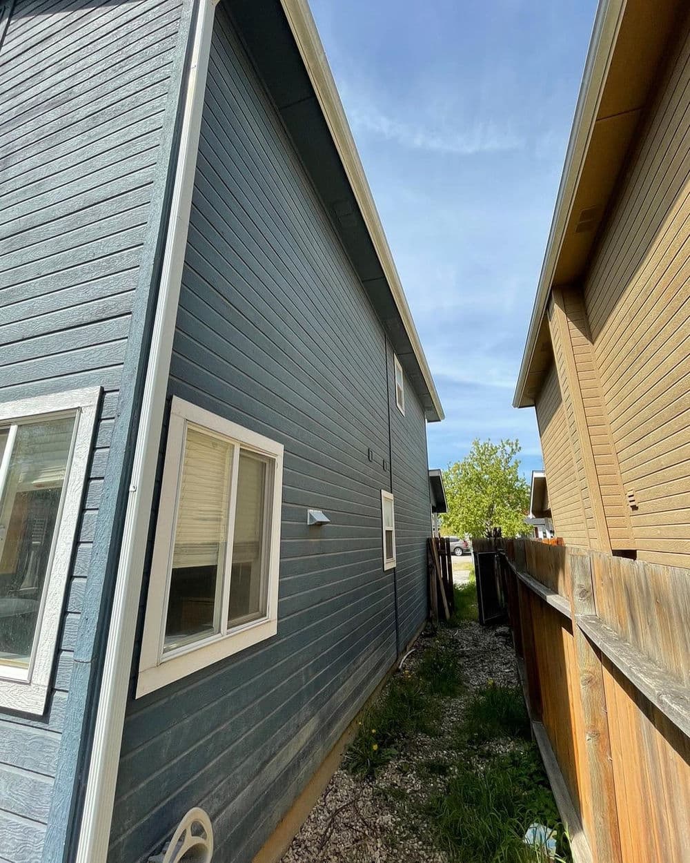 Side view of a blue house with windows, narrow pathway, and wooden fence. Clear sky above.